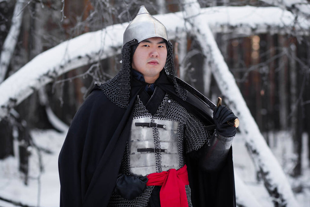   Portrait of a medieval warrior in armor, a helmet and a black cloak with a saber in his hands against the background of a winter forest and snow. - Фото, изображение