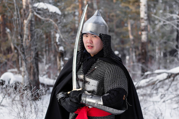   Portrait of a medieval warrior in armor, a helmet and a black cloak with a saber in his hands against the background of a winter forest and snow. - Фото, изображение