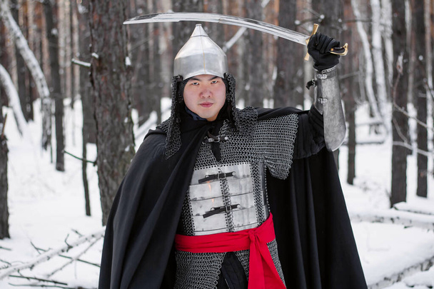   Retrato de un guerrero medieval con armadura, casco y manto negro con un sable en las manos sobre el fondo de un bosque invernal y nieve
. - Foto, imagen