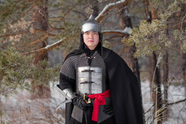   Retrato de um guerreiro medieval em armadura, um capacete e um manto preto com um sabre nas mãos contra o fundo de uma floresta de inverno e neve
. - Foto, Imagem
