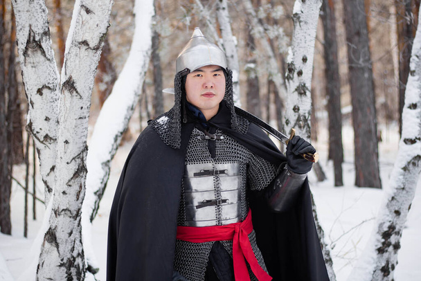   Egy középkori harcos portréja páncélban, sisakkal és fekete köpennyel a kezében, karddal a kezében, a téli erdő és a hó hátterében.. - Fotó, kép