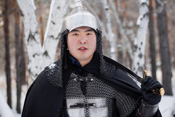  鎧を着た中世の戦士の肖像画、冬の森と雪を背景に彼の手にサーベルとヘルメットと黒のマント. - 写真・画像