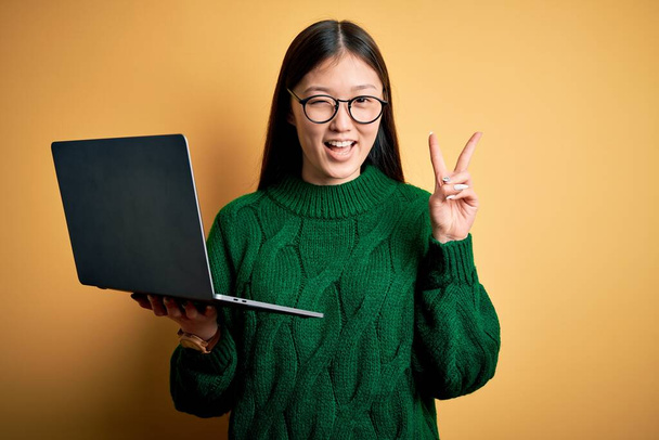 Fiatal ázsiai üzletasszony szemüveget visel, és számítógépes laptoppal dolgozik, boldog arccal mosolyog a kamerára, győzelmi táblát téve az ujjaival. Második.. - Fotó, kép