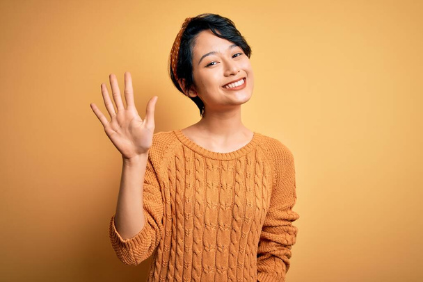 Молодая красивая азиатская девушка в обычном свитере и диадеме стоит на желтом фоне и показывает пальцами номер пять, улыбаясь уверенно и счастливо
. - Фото, изображение