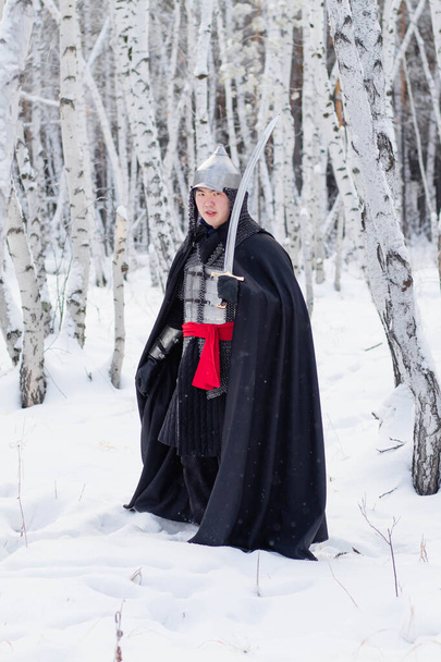   Guerreiro medieval em cadeia de correio, capacete, manto preto com um sabre nas mãos contra o fundo de uma floresta de bétula de inverno
. - Foto, Imagem