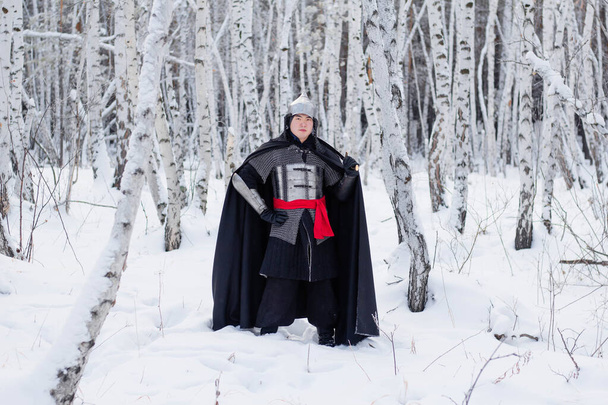   Medieval warrior in chain mail, helmet, black cloak with a saber in his hands against the background of a winter birch forest. - Φωτογραφία, εικόνα