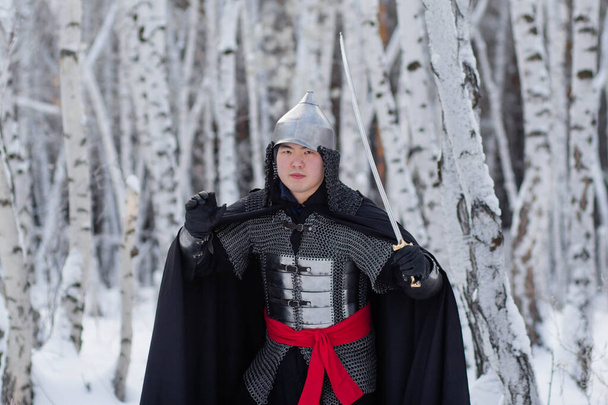  Guerrier médiéval dans la chaîne de courrier, casque, manteau noir avec un sabre dans ses mains sur le fond d'une forêt de bouleaux d'hiver
. - Photo, image