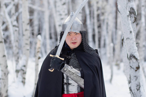   Středověký válečník v kroužkové zbroji, helmě, černém plášti s šavlí v rukou na pozadí zimního břízového lesa. - Fotografie, Obrázek