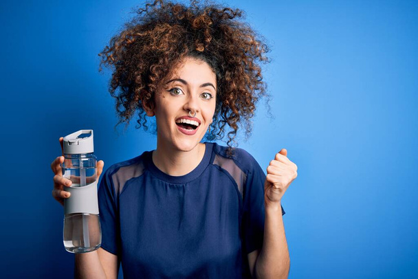 Young beautiful sporty woman with curly hair and piercing doing sport holding bottle of water screaming proud and celebrating victory and success very excited, cheering emotion - Photo, image