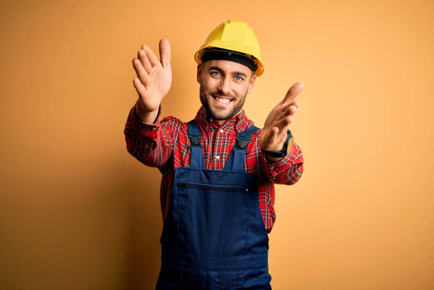 Молодой строитель в строительной форме и защитном шлеме на жёлтом изолированном фоне смотрит на камеру, улыбаясь с распростертыми объятиями. Веселое выражение, охватывающее счастье
. - Фото, изображение