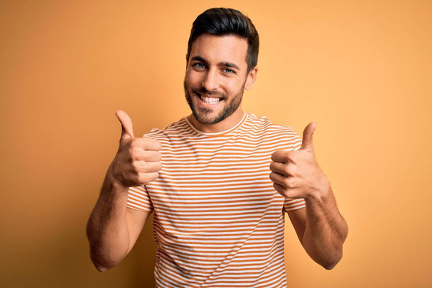 Young handsome man with beard wearing casual striped t-shirt over yellow background success sign doing positive gesture with hand, thumbs up smiling and happy. Cheerful expression and winner gesture. - Photo, Image