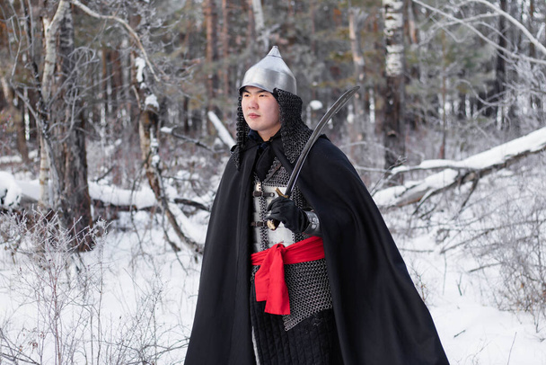Medieval warrior in armor, helmet, black cloak with a saber in his hands walking in the winter in the forest. - Zdjęcie, obraz