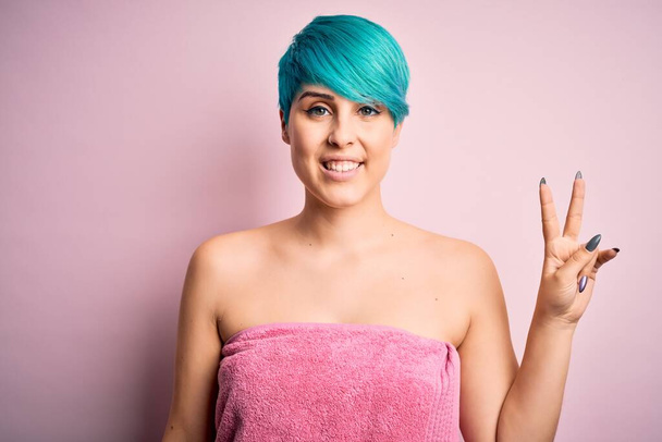 Giovane donna con i capelli blu moda indossa asciugamano dopo la doccia su sfondo rosa sorridente con viso felice ammiccando alla fotocamera facendo segno di vittoria. Numero due.
. - Foto, immagini