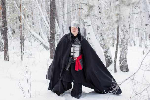 Medieval warrior in armor, helmet, black cloak with a saber in his hands walking in the winter in the forest. - Foto, afbeelding