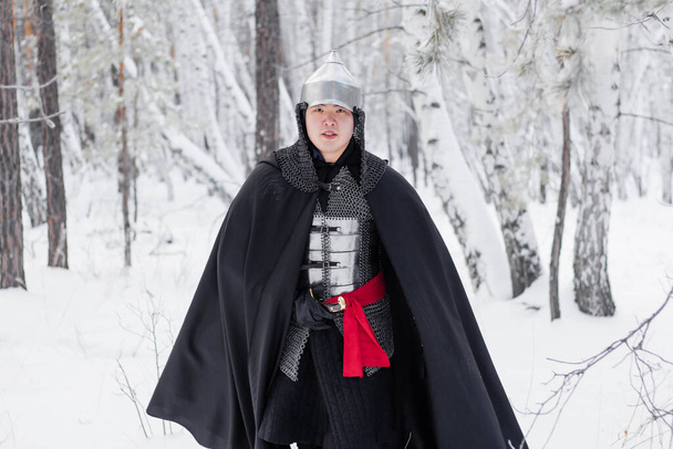 Medieval warrior in armor, helmet, black cloak with a saber in his hands walking in the winter in the forest. - Foto, immagini