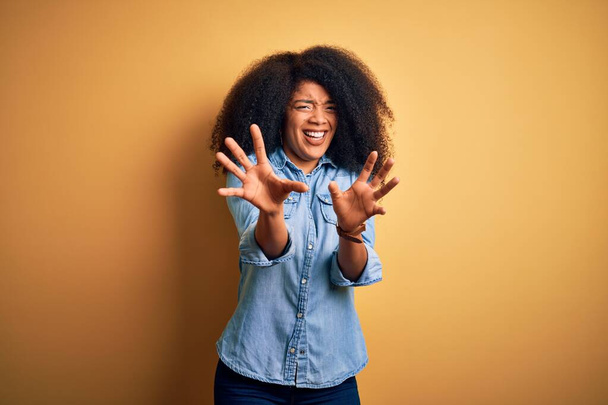 黄色の隔離された背景の上に立っているアフロの髪を持つ若い美しいアフリカ系アメリカ人女性は、ショックで叫んで、手で恐怖表現を停止ジェスチャーで恐れています。パニック概念. - 写真・画像