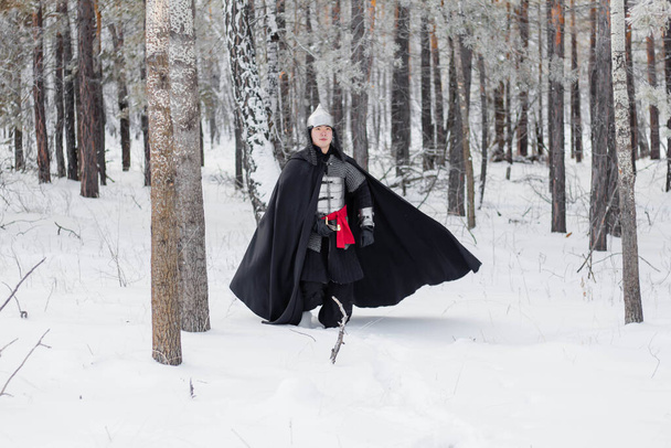 Középkori harcos páncélban, sisakban, fekete köpenyben, karddal a kezében, télen sétálva az erdőben.. - Fotó, kép