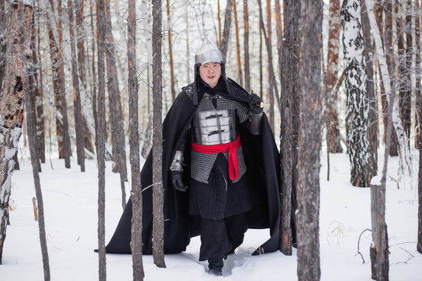 Średniowieczny wojownik w zbroi, hełmie, czarnym płaszczu z szablą w rękach, chodzący zimą po lesie.. - Zdjęcie, obraz