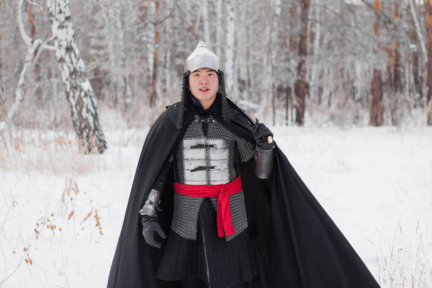 Guerrero medieval en armadura, casco, manto negro con un sable en las manos caminando en el invierno en el bosque
. - Foto, Imagen