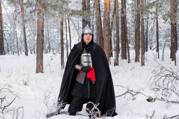Medieval warrior in armor, helmet, black cloak with a saber in his hands walking in the winter in the forest. - Foto, afbeelding