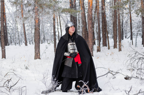 Medieval warrior in armor, helmet, black cloak with a saber in his hands walking in the winter in the forest. - Fotografie, Obrázek