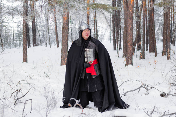 Medieval warrior in armor, helmet, black cloak with a saber in his hands walking in the winter in the forest. - Photo, Image