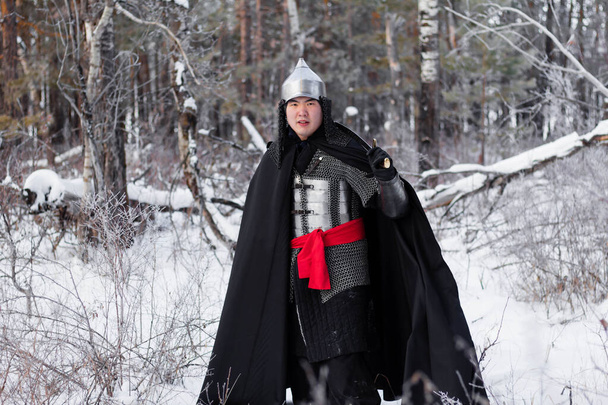 Medieval warrior in armor, helmet, black cloak with a saber in his hands walking in the winter in the forest. - Foto, Imagen