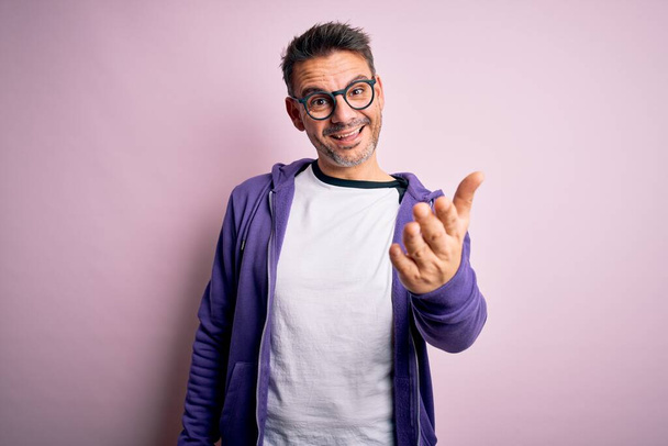 Young handsome man wearing purple sweatshirt and glasses standing over pink background smiling friendly offering handshake as greeting and welcoming. Successful business. - Photo, Image
