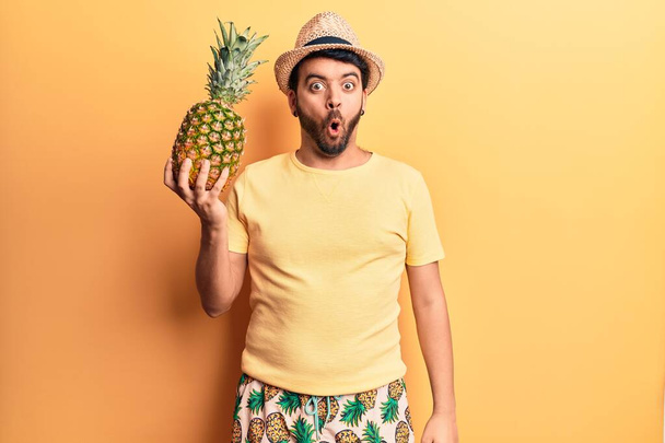 Junger hispanischer Mann in Badebekleidung und Sommermütze mit Ananas in der Hand, erschrocken und erstaunt mit offenem Mund für Überraschung, ungläubiges Gesicht  - Foto, Bild