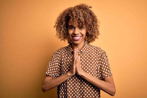 Joven mujer afroamericana hermosa vistiendo camisa casual de pie sobre fondo amarillo rezando con las manos juntas pidiendo perdón sonriendo confiado
. - Foto, Imagen