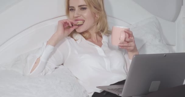 Freelancer working on laptop eating cookie and enjoys a cup of hot cocoa or tea while long working night. Business woman relaxes in bed after a long flight. Business concept. Prores 422 - Filmmaterial, Video
