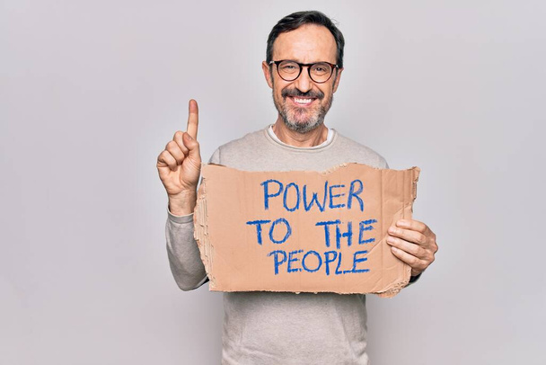 Mann mittleren Alters, der soziale Rechte fordert, hält Banner mit Macht an das Volk, lächelt mit einer Idee oder einer Frage und zeigt mit glücklichem Gesicht auf den Finger, Nummer eins - Foto, Bild