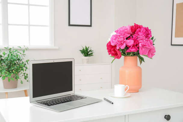 Vase with beautiful peony flowers and laptop on table in room - 写真・画像