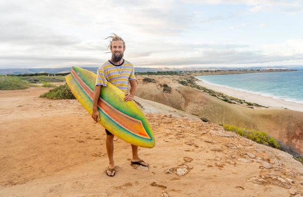 Atractivo surfista hipster rubia australiana con rastas y barba mirando el mar celebración de tabla de surf vintage fresco al atardecer. Volver a surfear, Deportes al aire libre estilo de vida de aventura en la Nueva Normal. - Foto, imagen