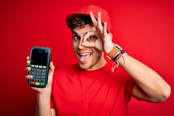 Nuori vaalea mies kihara tukka yllään korkki tilalla dataphone yli punainen tausta onnellinen kasvot hymyillen tekee ok merkki käsin katsellen sormien läpi - Valokuva, kuva