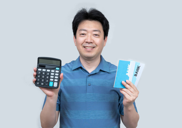 Azjata w średnim wieku trzymający w ręku paszport bankowy i kalkulator na szarym tle. - Zdjęcie, obraz