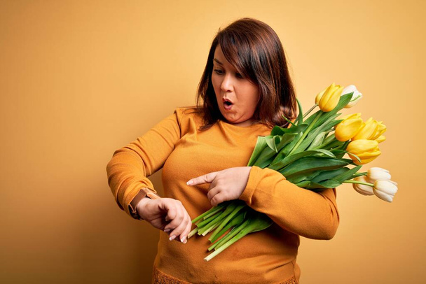 Красивая плюс размер женщина держит романтический букет натуральных цветов тюльпанов на желтом фоне, глядя на часы время беспокоится, боится опоздать
 - Фото, изображение