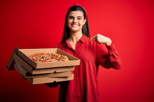 Young beautiful brunette woman holding delivery boxes with Italian pizza over red background with surprise face pointing finger to himself - Foto, afbeelding