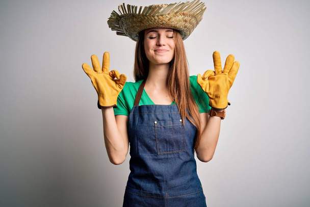 Young beautiful redhead farmer woman wearing apron and hat over white background relax and smiling with eyes closed doing meditation gesture with fingers. Yoga concept. - Photo, image