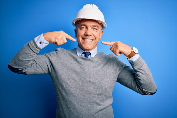 Keski-ikä komea harmaa tukka insinööri mies yllään turvakypärä yli sininen tausta hymyilevä iloinen osoittaa ja osoittaa sormin hampaat ja suu. Hammasterveyden käsite. - Valokuva, kuva