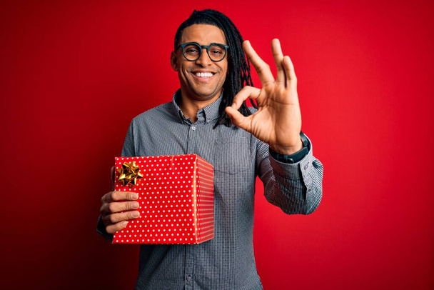 Афроамериканец с дредами держит настоящую коробку на красном изолированном фоне делает ОК знак с пальцами, отличный символ
 - Фото, изображение