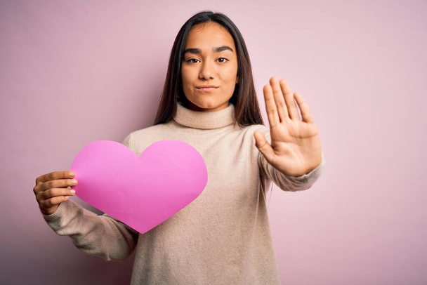 Fiatal gyönyörű ázsiai nő kezében rózsaszín szív áll az elszigetelt háttér nyitott kézzel csinál stop jel komoly és magabiztos kifejezés, védelmi gesztus - Fotó, kép
