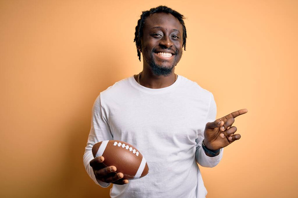 若いですアフリカ人選手男遊びラグビー保持アメリカンフットボールボール上の黄色の背景非常に幸せなポインティングで手と指でサイド - 写真・画像
