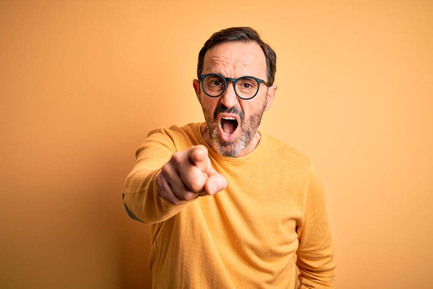 Mann mittleren Alters in lässigem Pullover und Brille vor isoliertem gelben Hintergrund, der unzufrieden und frustriert in die Kamera zeigt, wütend und wütend auf dich - Foto, Bild