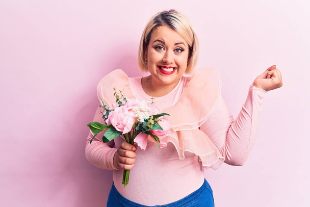 Schöne blonde Plus-Size-Frau mit einem Strauß pinkfarbener Blumen vor isoliertem Hintergrund, die stolz schreit und Sieg und Erfolg mit erhobenem Arm feiert - Foto, Bild