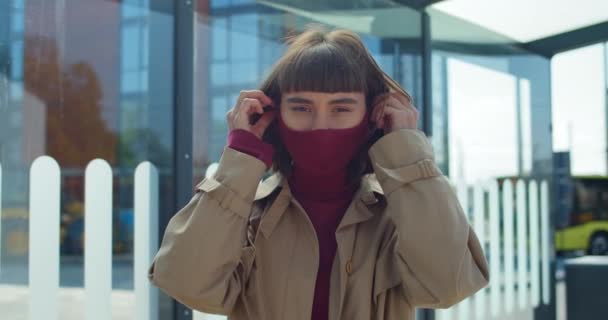 Portrait of millennial girl wearing protective cotton mask and looking to camera.Close up view of young woman standing at public transport stop. Concept of virus protection. - Filmati, video