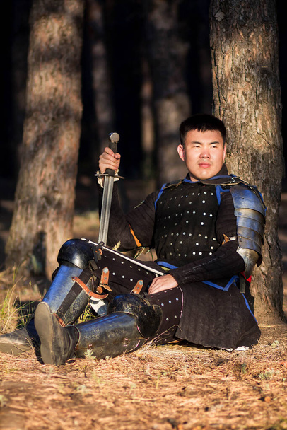A warrior in black armor with a sword in his hands sits on the ground, leaning against a tree against the backdrop of a gloomy forest. - Foto, Imagem