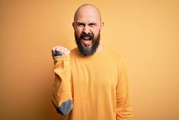 Красивый лысый мужчина с бородой в случайном свитере стоит на жёлтом фоне злой и безумный поднимающий кулак расстроенный и разъяренный, крича от ярости. Ярость и агрессивность
. - Фото, изображение