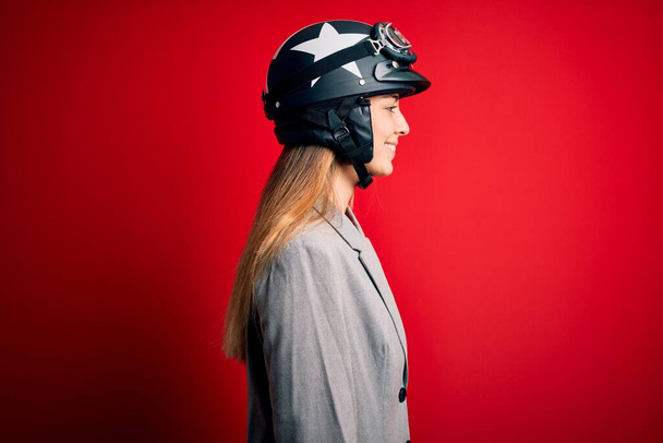 Jovem mulher motociclista loira bonita usando capacete de motocicleta sobre fundo vermelho olhando para o lado, relaxar pose perfil com rosto natural com sorriso confiante
. - Foto, Imagem