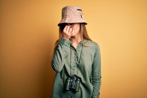 Όμορφη ξανθιά εξερευνήτρια γυναίκα με μπλε μάτια φορώντας καπέλο και γυαλιά χρησιμοποιώντας κιάλια κουρασμένο τρίψιμο μύτη και τα μάτια αίσθημα κόπωσης και πονοκέφαλο. Έννοια άγχους και απογοήτευσης. - Φωτογραφία, εικόνα